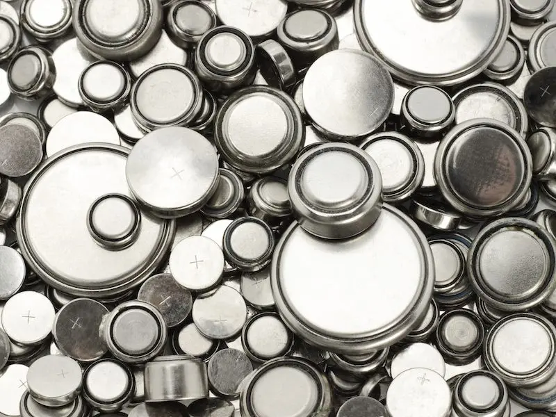 A close-up view of a pile of various sizes of round, silver-colored button cell batteries, some marked with a plus sign on top.