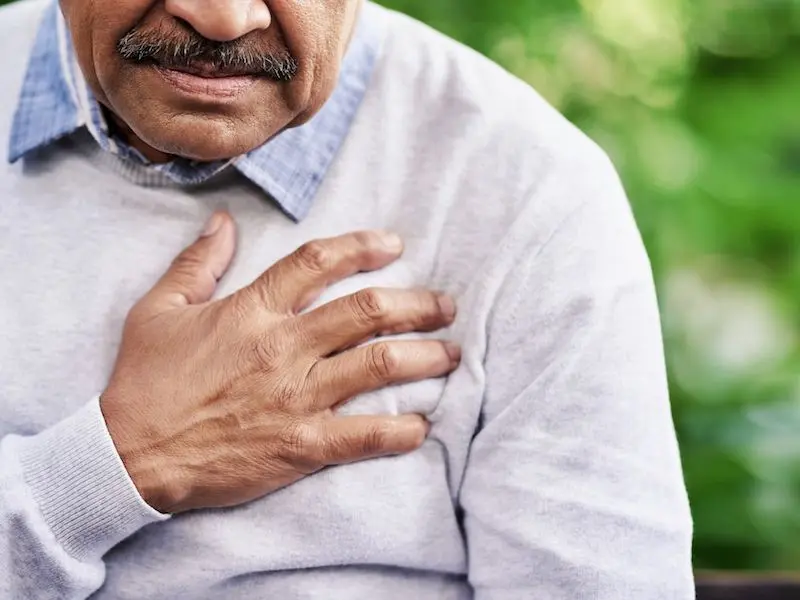 An older man wearing a light gray sweater and blue collared shirt holds his hand to his chest, suggesting he may be experiencing chest pain or discomfort, with a blurred green background.