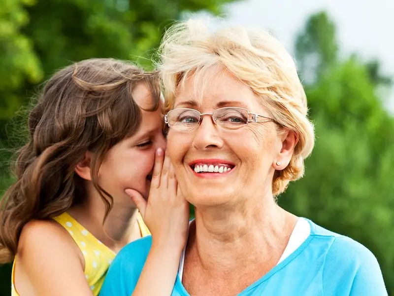 A smiling older woman with glasses stands outdoors while a young girl whispers in her ear. The background is green and blurred, suggesting a park or garden setting. Both appear happy and close.