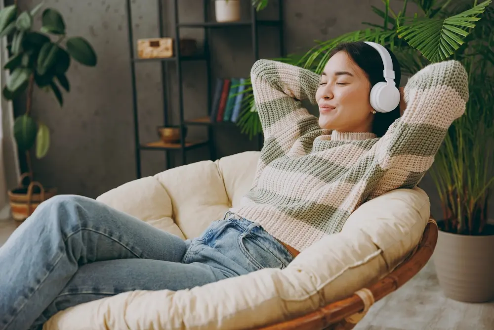 A woman sits in a cozy chair with her arms behind her head, eyes closed, and a relaxed smile, wearing headphones. She is dressed in a striped sweater and jeans, surrounded by plants and bookshelves.