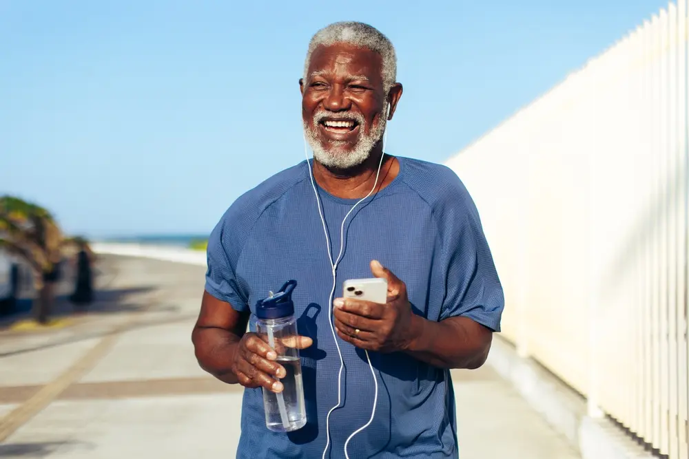 Older man with gray hair and beard smiling while holding a water bottle and smartphone, wearing headphones and a blue shirt, walking outdoors along a sunny pathway near a fence.
