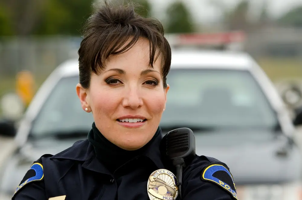 A female police officer stands confidently with arms crossed, smiling in front of a police car. She wears a uniform, badge, and shoulder radio, with short hair and blurred trees in the background.