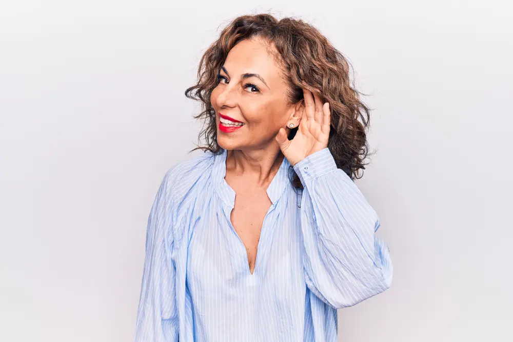 Smiling woman with curly hair wearing a light blue shirt holds her hand to her ear, as if trying to hear better, while standing against a plain white background.
