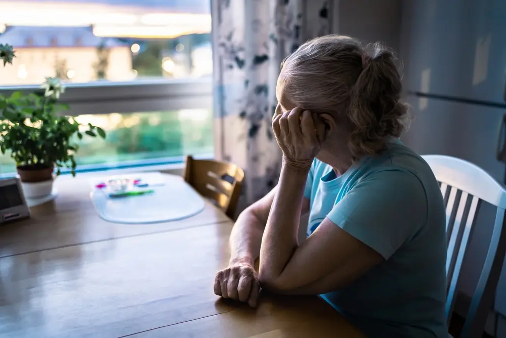 An elderly woman with gray hair sits at a wooden table, resting her head on her hand and looking out a window. A plant and some items are on the table, and the room appears dimly lit.