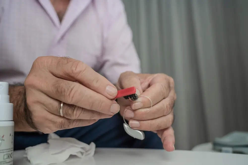 A person wearing a pink checkered shirt and a wedding ring uses a small red tool to clean or adjust a hearing aid, with a white glove and bottle nearby on a white surface.