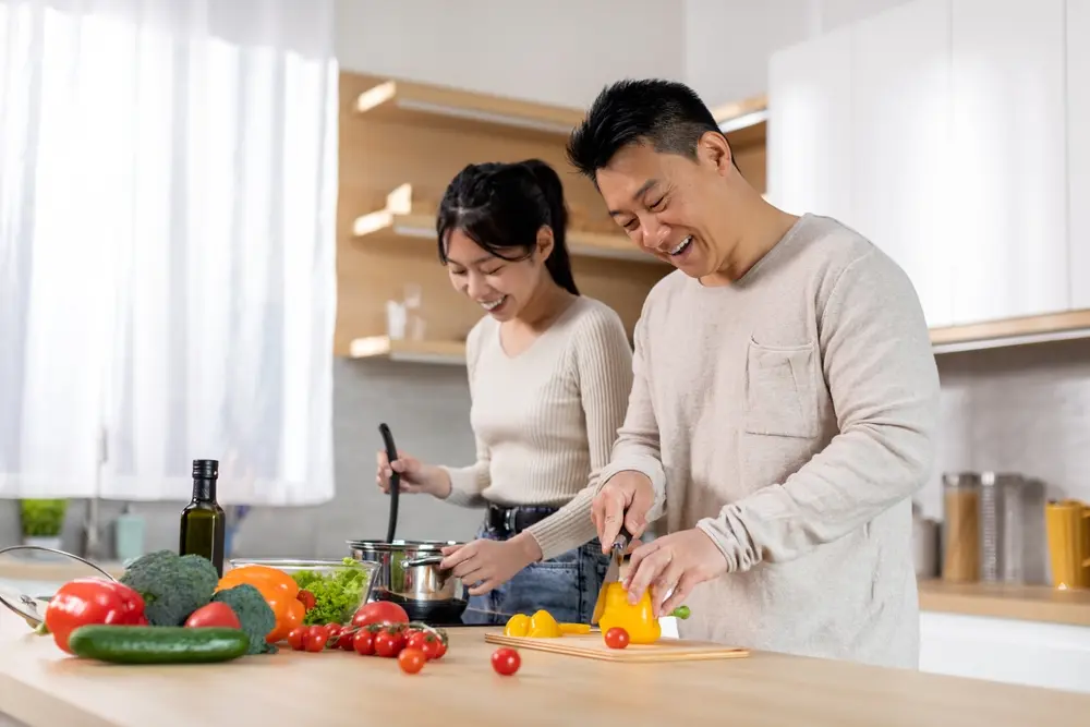 A man and woman preparing food together in a bright kitchen; he slices a yellow bell pepper while she cooks at the stove. Fresh vegetables are spread on the counter in front of them.
