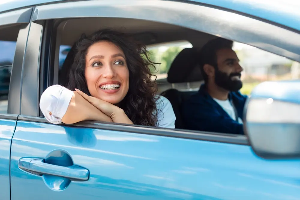 A smiling woman with braces leans out the window of a blue car, resting her arms on the door, while a man sits in the drivers seat, both appearing happy and relaxed.