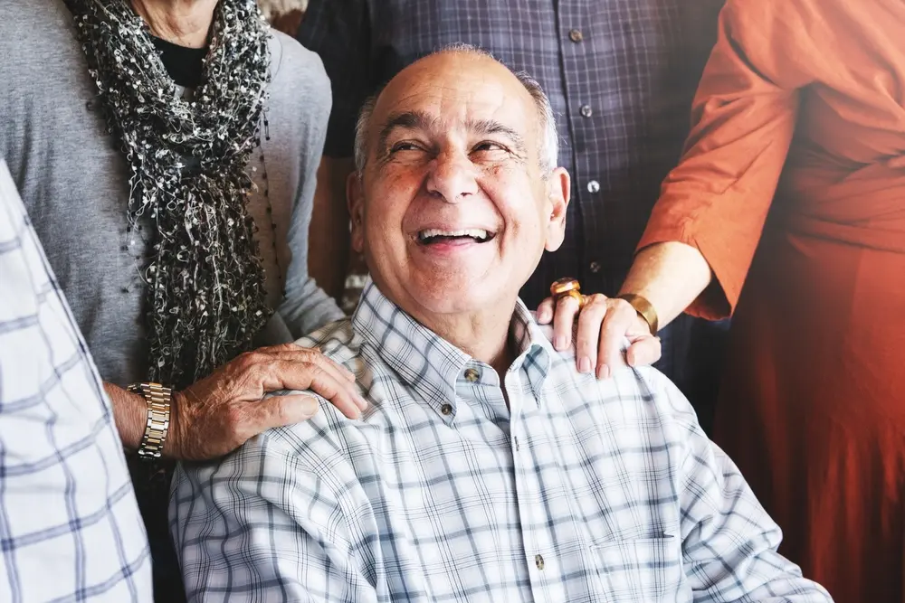 An older man in a plaid shirt smiles joyfully while sitting, surrounded by friends whose hands rest supportively on his shoulders. Warm, natural light shines on the group, creating a cheerful and friendly atmosphere.