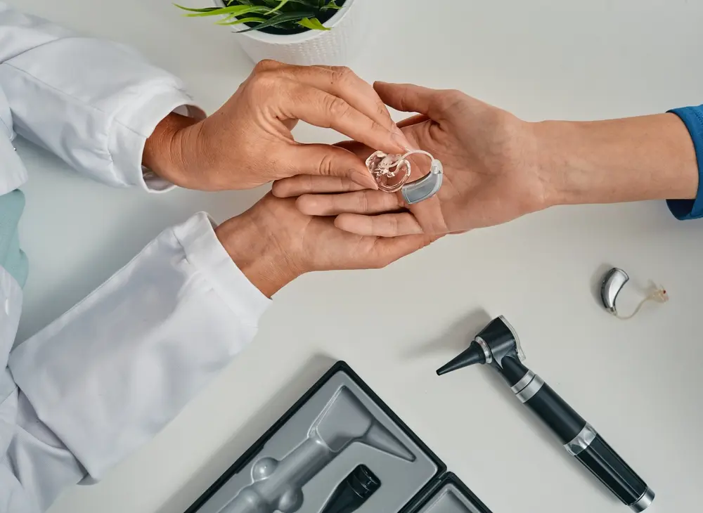 A doctor hands a hearing aid to a patient across a white table with an otoscope, another hearing aid, and a small plant visible nearby.