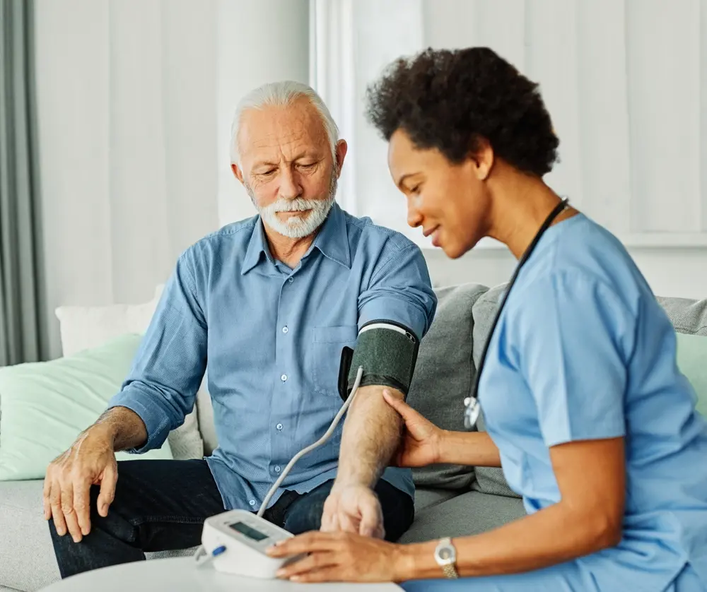 A nurse in blue scrubs checks the blood pressure of an elderly man with white hair and a mustache, who is seated on a couch at home. The nurse is operating a digital blood pressure monitor.