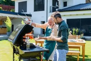 Two men smiling and grilling food together on a barbecue in a backyard, with a modern house and outdoor dining table in the background, on a sunny day.