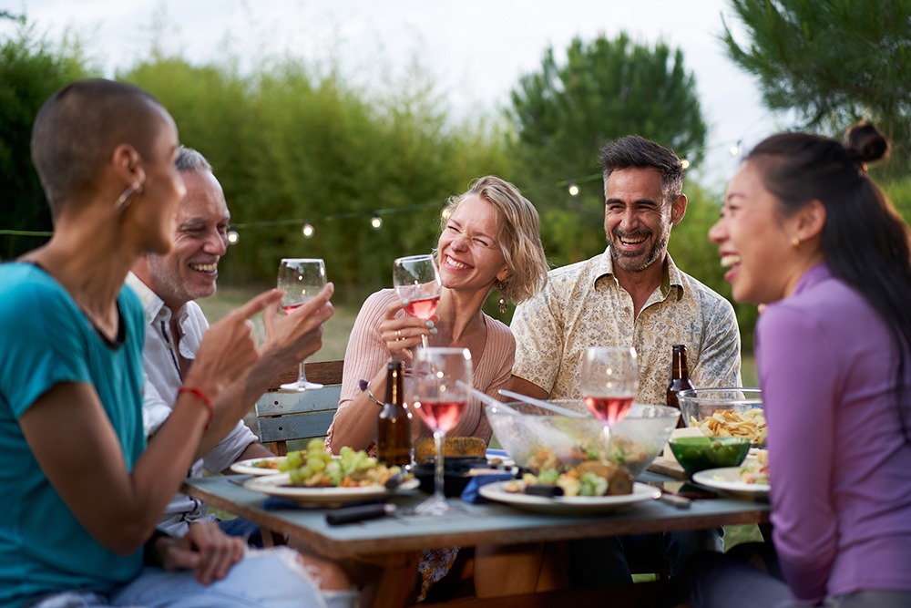 Five people sit around an outdoor table, laughing and holding glasses of wine, with plates of food in front of them and string lights in the background. The mood is joyful and relaxed.