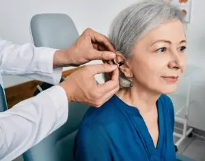 An audiologist fits a hearing aid on an older woman with gray hair, who is seated and smiling, wearing a blue shirt in a medical office.