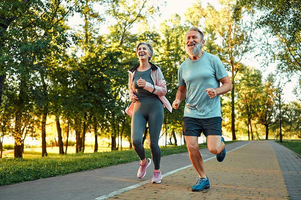 An older man and woman jog together on a paved path in a park, surrounded by green trees and sunlight. They are both smiling and appear to be enjoying their outdoor exercise.
