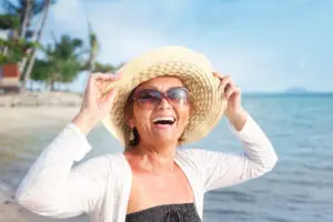 Smiling woman wearing sunglasses and a wide-brimmed hat stands on a sunny beach, with the sea and palm trees in the background. She is holding her hat and enjoying the warm weather.