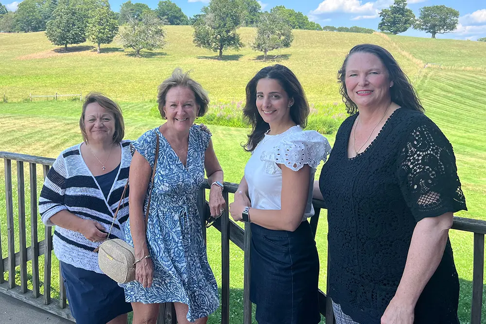 Four women stand on a wooden deck in front of a scenic green field with trees, smiling at the camera on a sunny day. They are dressed in summer clothing.